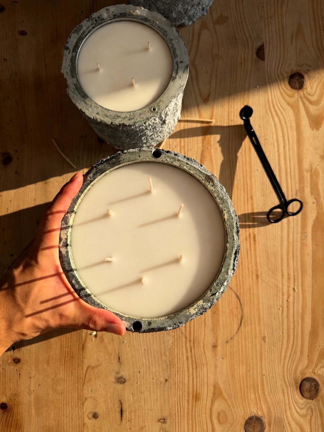 A hand holding a large multi-wick candle poured into a rough, sea-worn vessel, seen from above in sunlight.